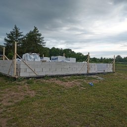 WIKO BOUW Grzegorz Serafin - Wznoszenie ścian z białych bloczków Solbet na fundamencie. Widoczne drewniane podpory i zapas materiału na paletach. W tle zielone drzewa i pochmurne niebo.