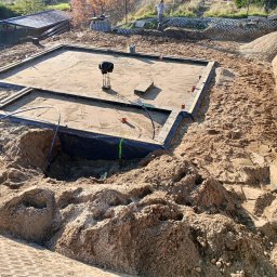 WIKO BOUW Grzegorz Serafin - Przygotowany teren pod fundamenty z widoczną izolacją pionową i poziomą. Na piasku leżą narzędzia i materiały budowlane. W tle widać zieleń i elementy krajobrazu.