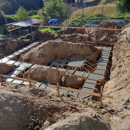 WIKO BOUW Grzegorz Serafin - Zalane betonem szalunki fundamentów domu w wykopie, widok z góry. Na pierwszym planie ziemia, w tle zieleń i zabudowania. Prace fundamentowe w toku.