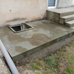 WIKO BOUW Grzegorz Serafin - Świeżo wylana, betonowa podstawa z otworem rewizyjnym i schodami wejściowymi do budynku. Widoczne ślady wilgoci na powierzchni betonu.