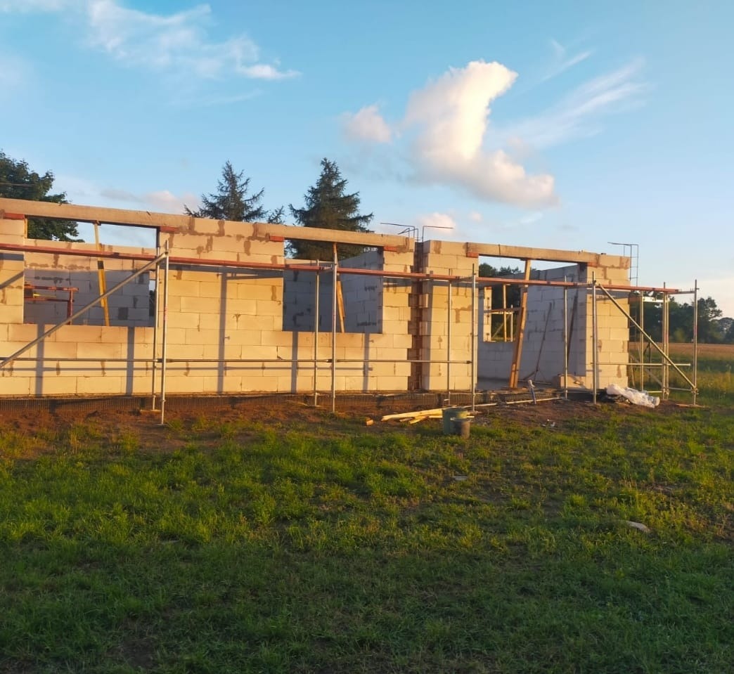 Wznoszenie ścian parterowego domu z bloczków, widoczne rusztowania i otwory okienne. Prace murarskie w trakcie realizacji na tle zielonej trawy i błękitnego nieba.