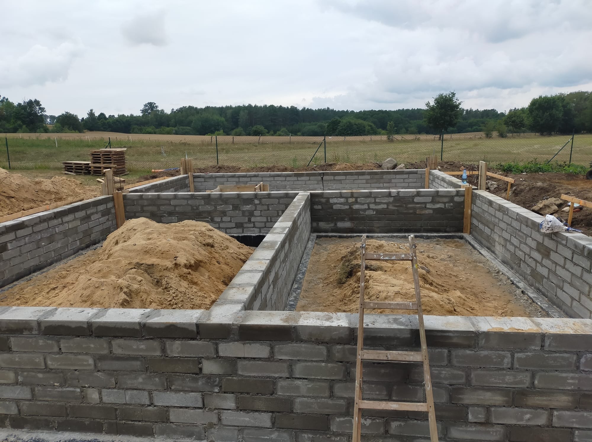 Wznoszenie fundamentów z bloczków betonowych, widok na częściowo zasypane piaskiem wnętrze, drabina oparta o ścianę, w tle łąka i las, pochmurne niebo.