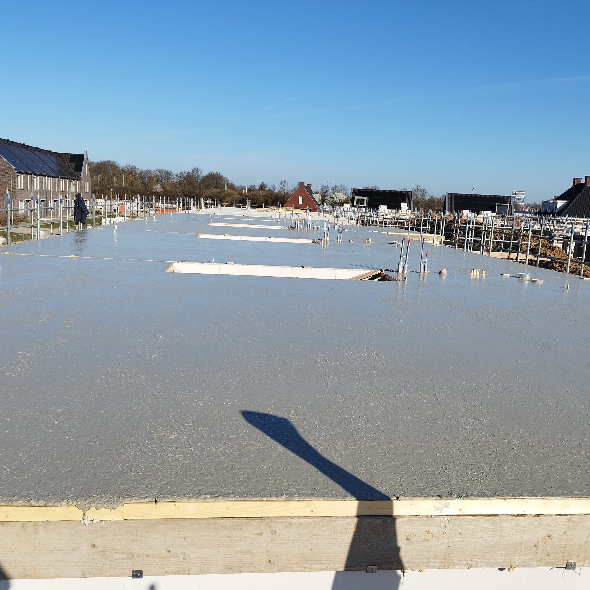 Wylany fundament z wystającymi rurami instalacyjnymi, widoczne szalunki i rusztowania. Nowe osiedle w tle pod błękitnym niebem.