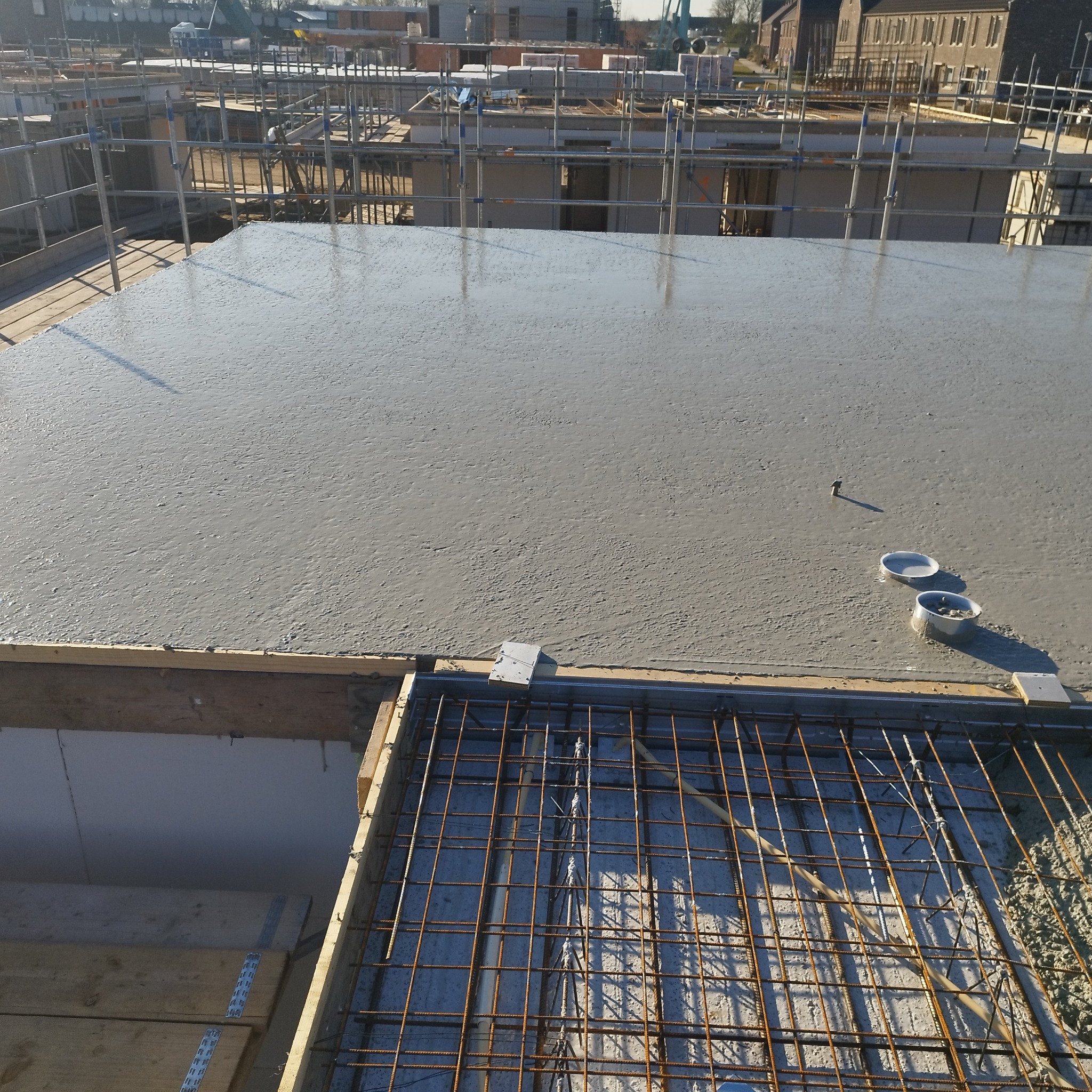 Zalany strop betonowy na budowie domu, widoczne stalowe zbrojenia i szalunki. W tle rusztowania i budynki w oddali. Jasne, naturalne światło.