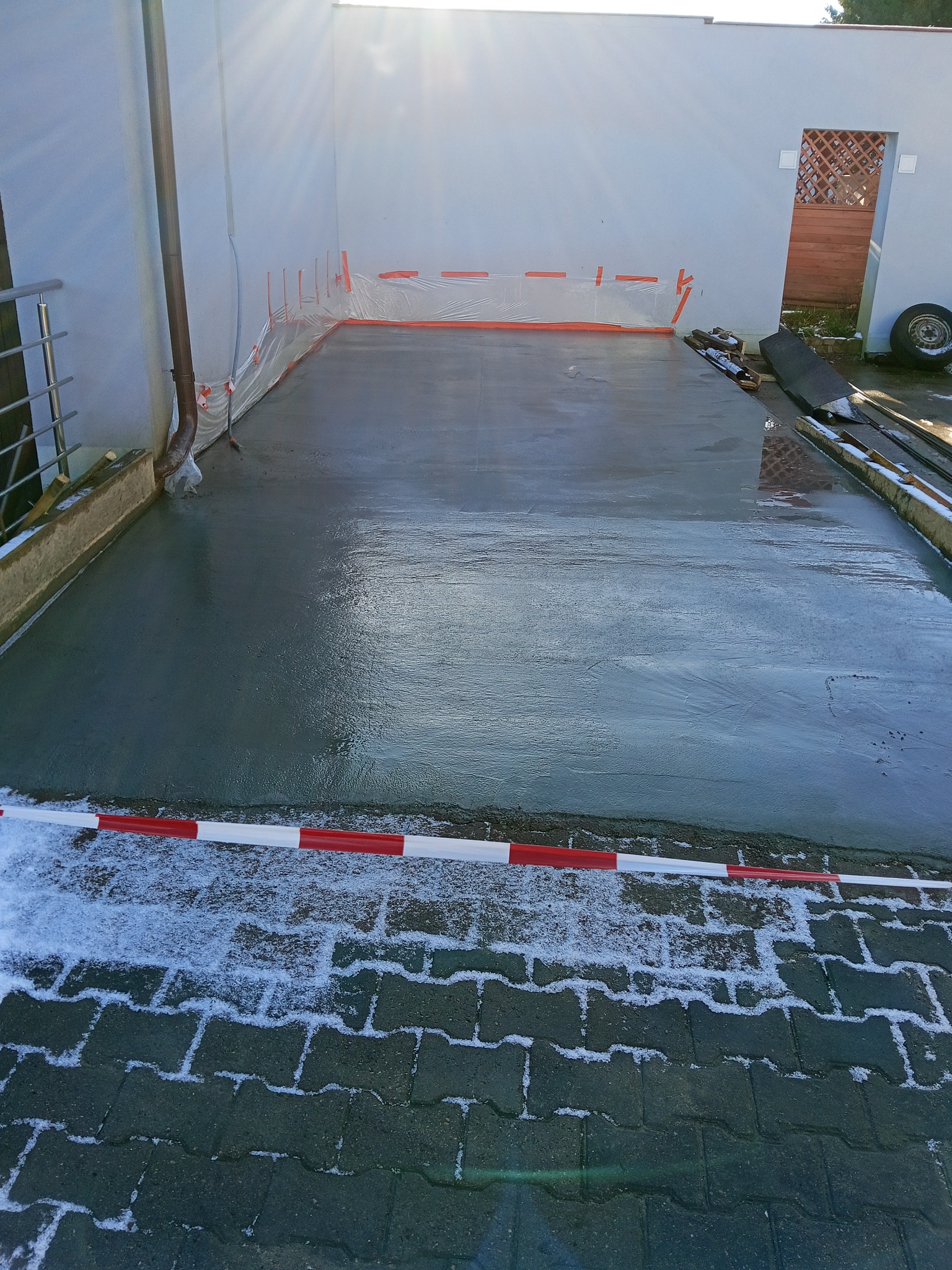 Świeżo wylana, gładka posadzka betonowa wewnątrz budynku, zabezpieczona taśmą ostrzegawczą. Widoczne ślady śniegu na kostce brukowej przed wejściem.