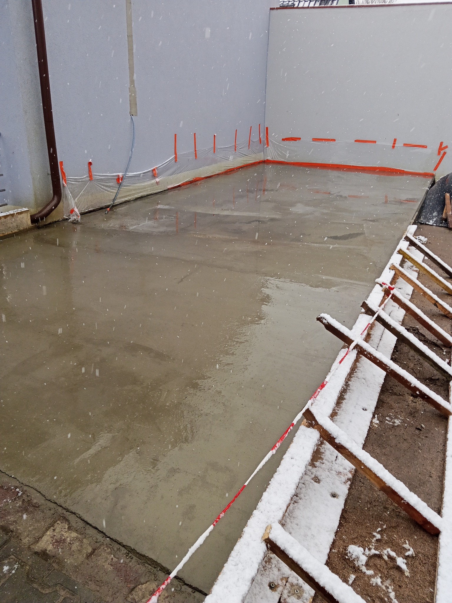 Świeżo wylana, mokra posadzka w trakcie opadów śniegu, zabezpieczona pomarańczową taśmą i folią. Widoczne drewniane elementy szalunku i fragment ściany budynku.