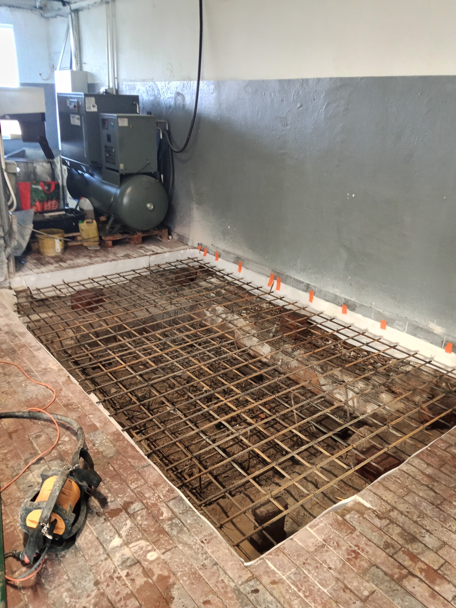 Zbrojenie pod wylewkę na podłodze z cegieł, widoczne pomarańczowe znaczniki na ścianie i sprzęt budowlany w tle. Przygotowanie do wylewki w pomieszczeniu.