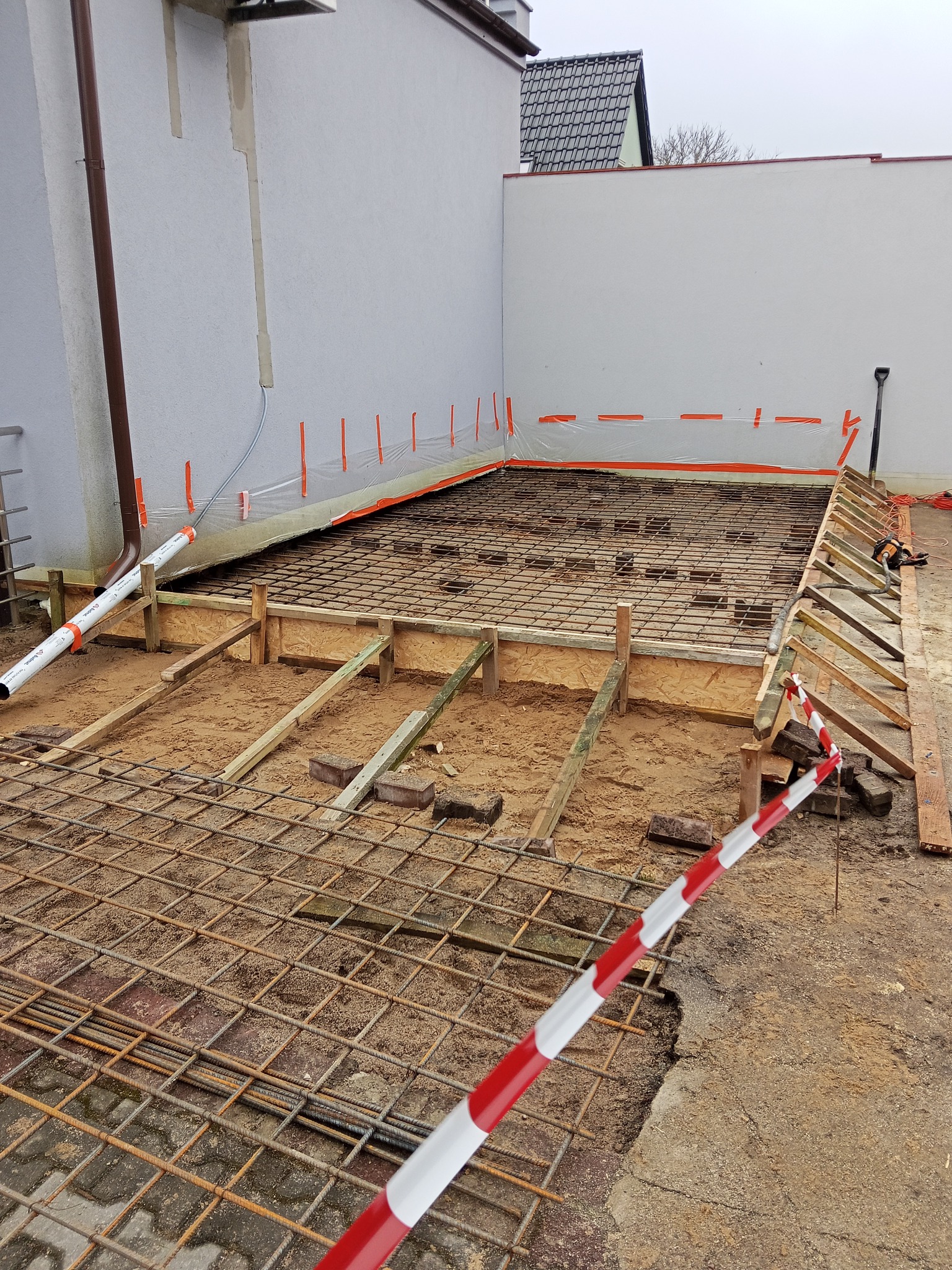 Zbrojenie fundamentu pod rozbudowę domu, widoczne drewniane szalunki i siatka zbrojeniowa. W tle ściana budynku i zabezpieczenia folią. Prace przygotowawcze do wylewki.