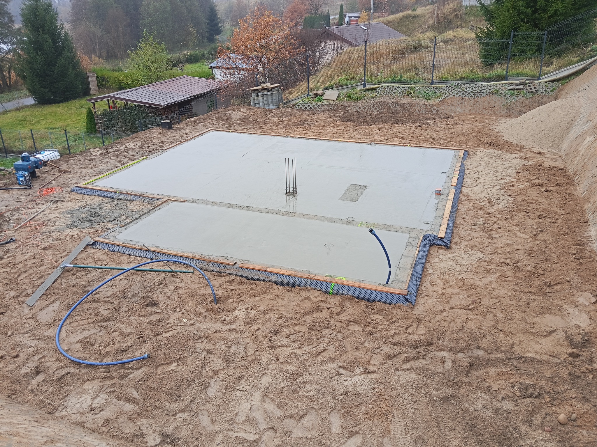 Wylany fundament pod budowę domu, widoczne wystające pręty zbrojeniowe. Świeży beton otoczony drewnianym szalunkiem i niebieskimi rurami. W tle zabudowa jednorodzinna.