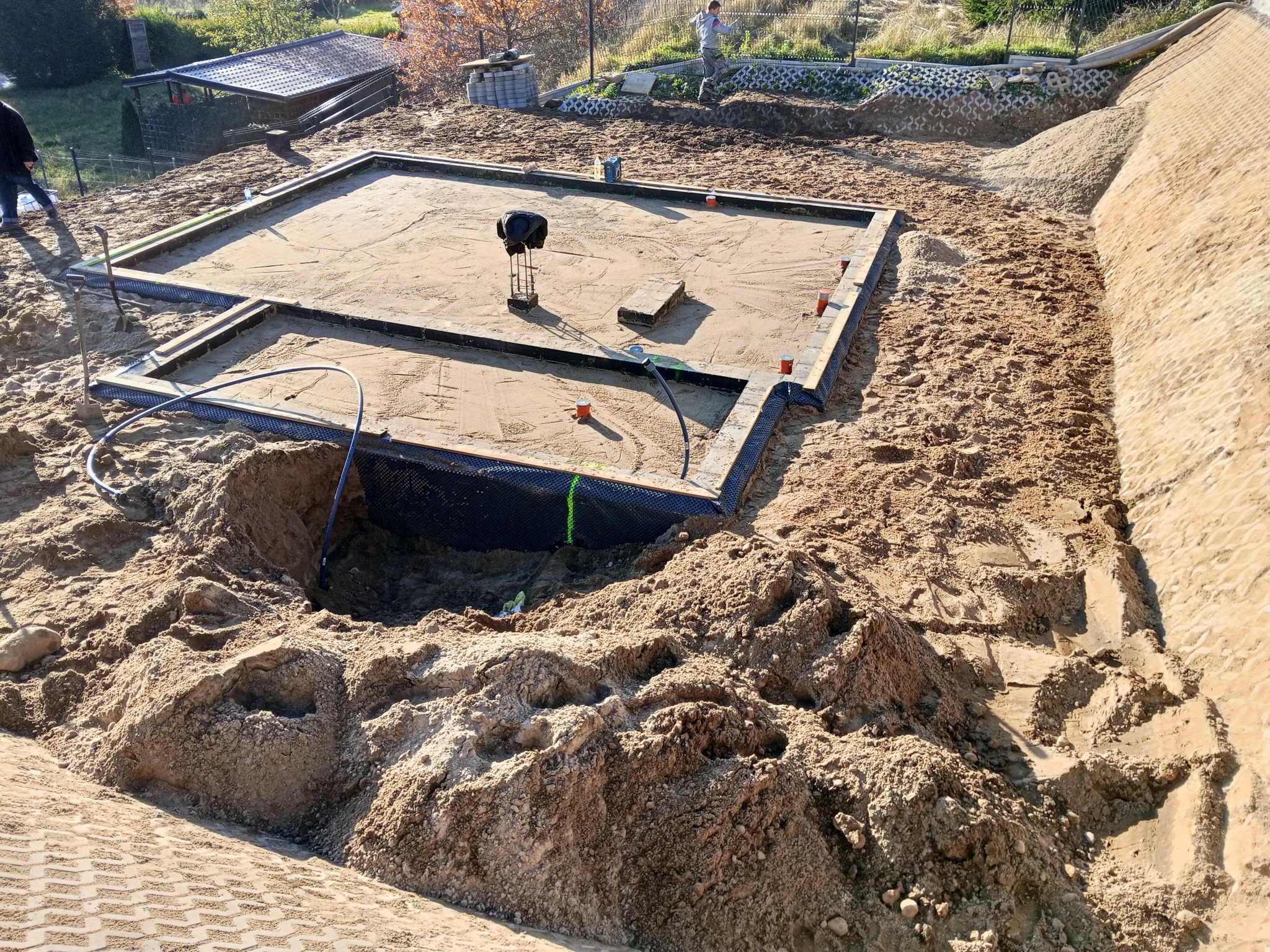 Przygotowany teren pod fundamenty z widoczną izolacją pionową i poziomą. Na piasku leżą narzędzia i materiały budowlane. W tle widać zieleń i elementy krajobrazu.