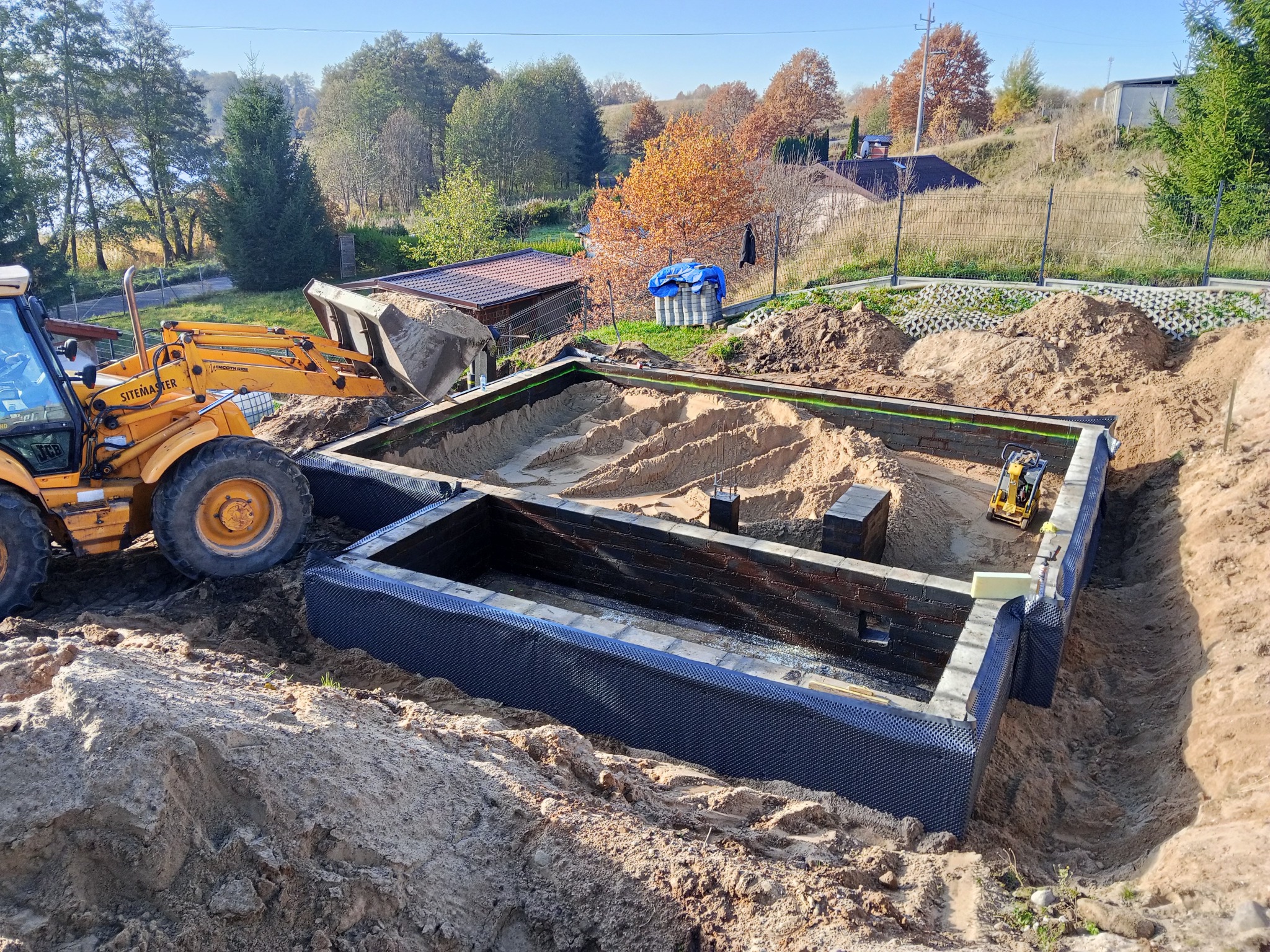 Wykop pod fundamenty z izolacją, częściowo zasypany piaskiem. Na pierwszym planie koparka, w tle zabudowa jednorodzinna i zieleń. Widok z góry, słoneczny dzień.