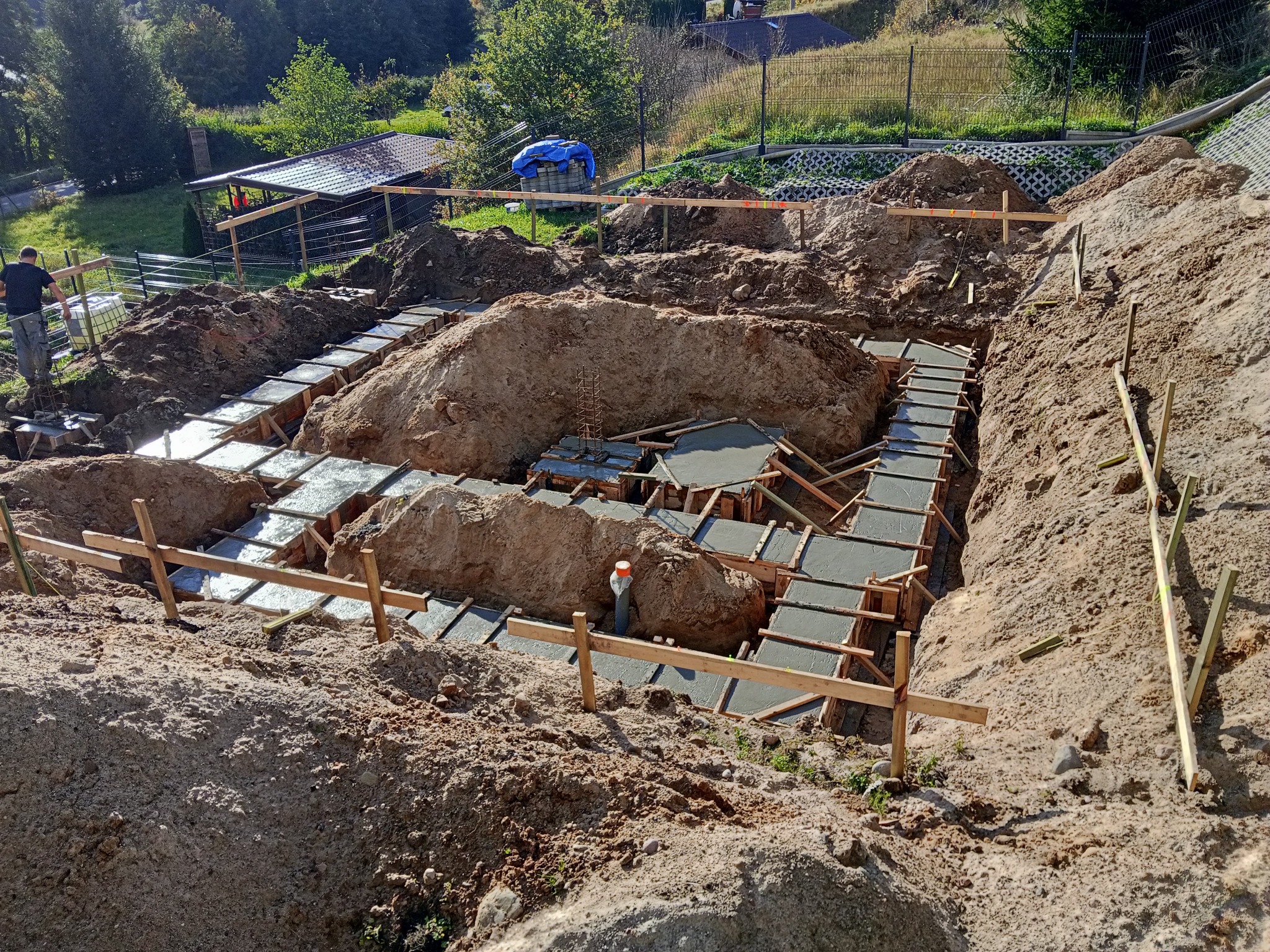 Zalane betonem szalunki fundamentów domu w wykopie, widok z góry. Na pierwszym planie ziemia, w tle zieleń i zabudowania. Prace fundamentowe w toku.