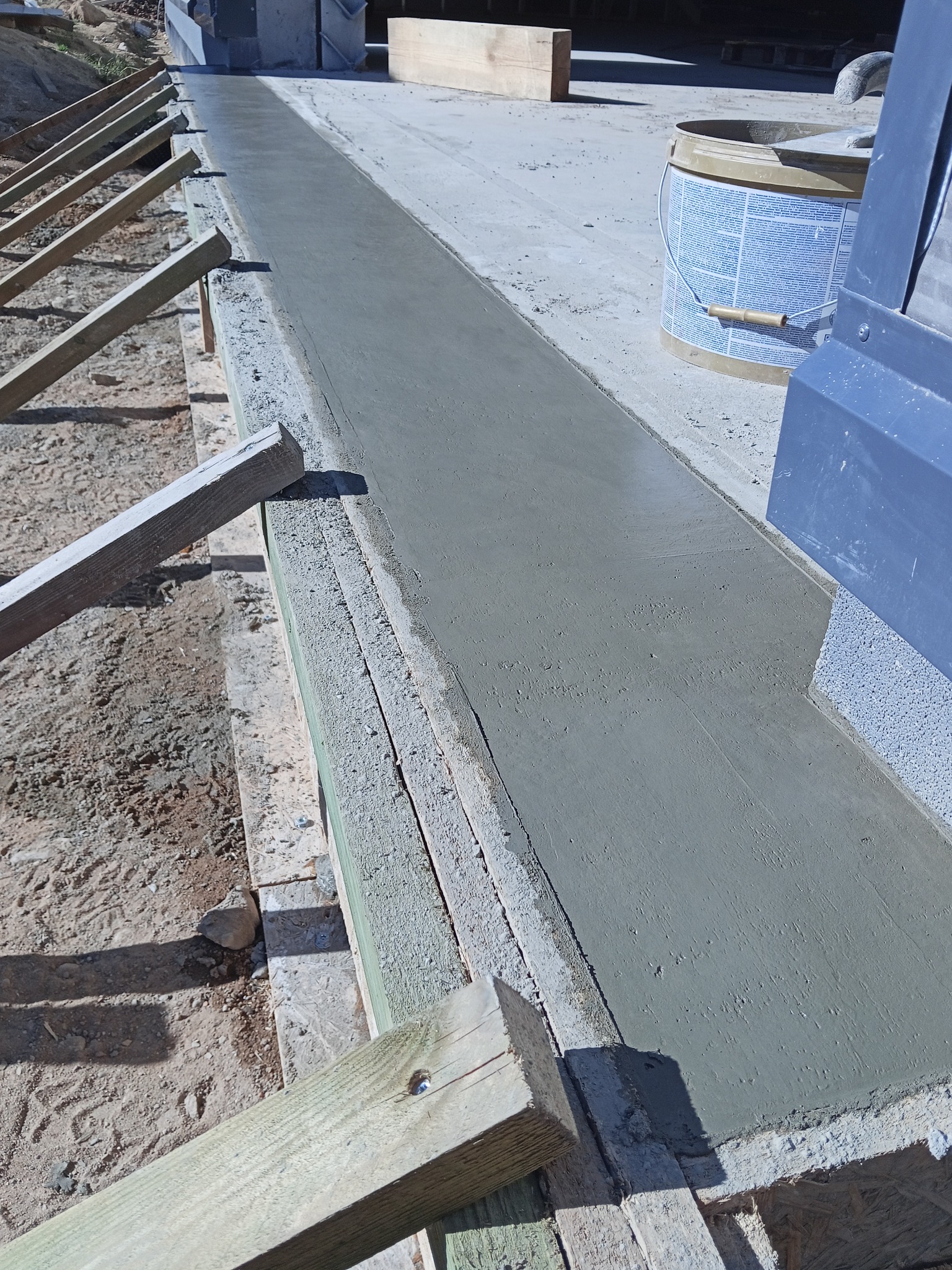 Świeżo wylany beton na wąskim fundamencie, widoczne drewniane szalunki i podpory. W tle wiadro z farbą i fragmenty konstrukcji.