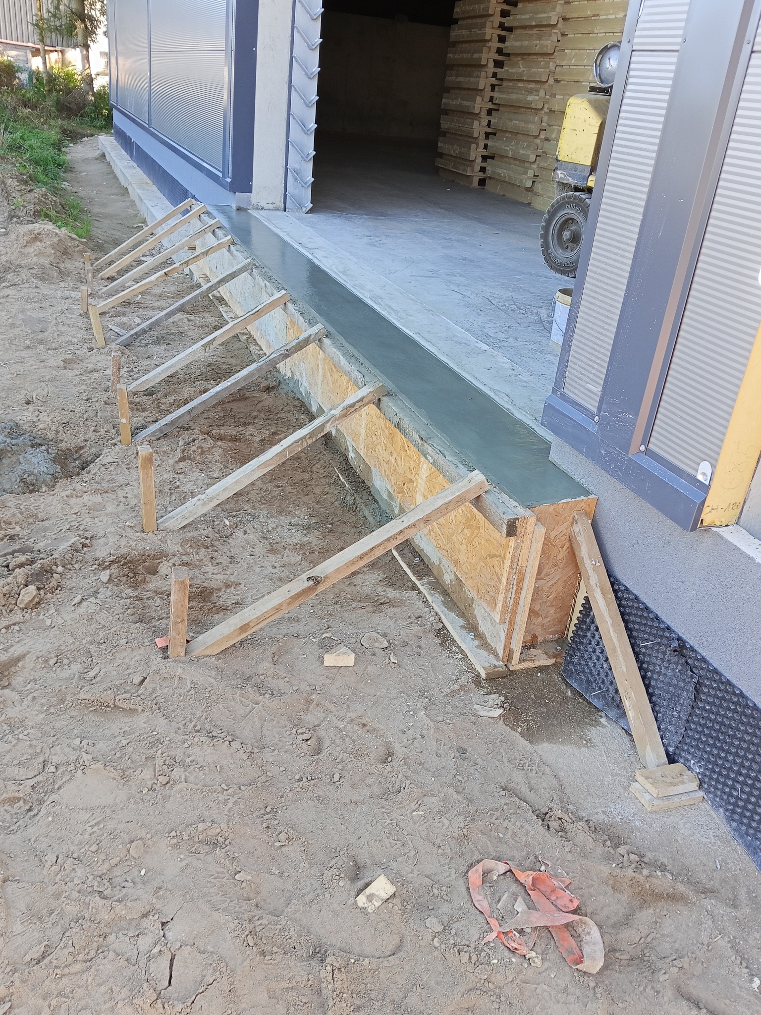 Wylewka betonowa przy budynku z szalunkiem z desek. Widoczna izolacja fundamentów i ziemia. Częściowo widoczny wózek widłowy w tle.