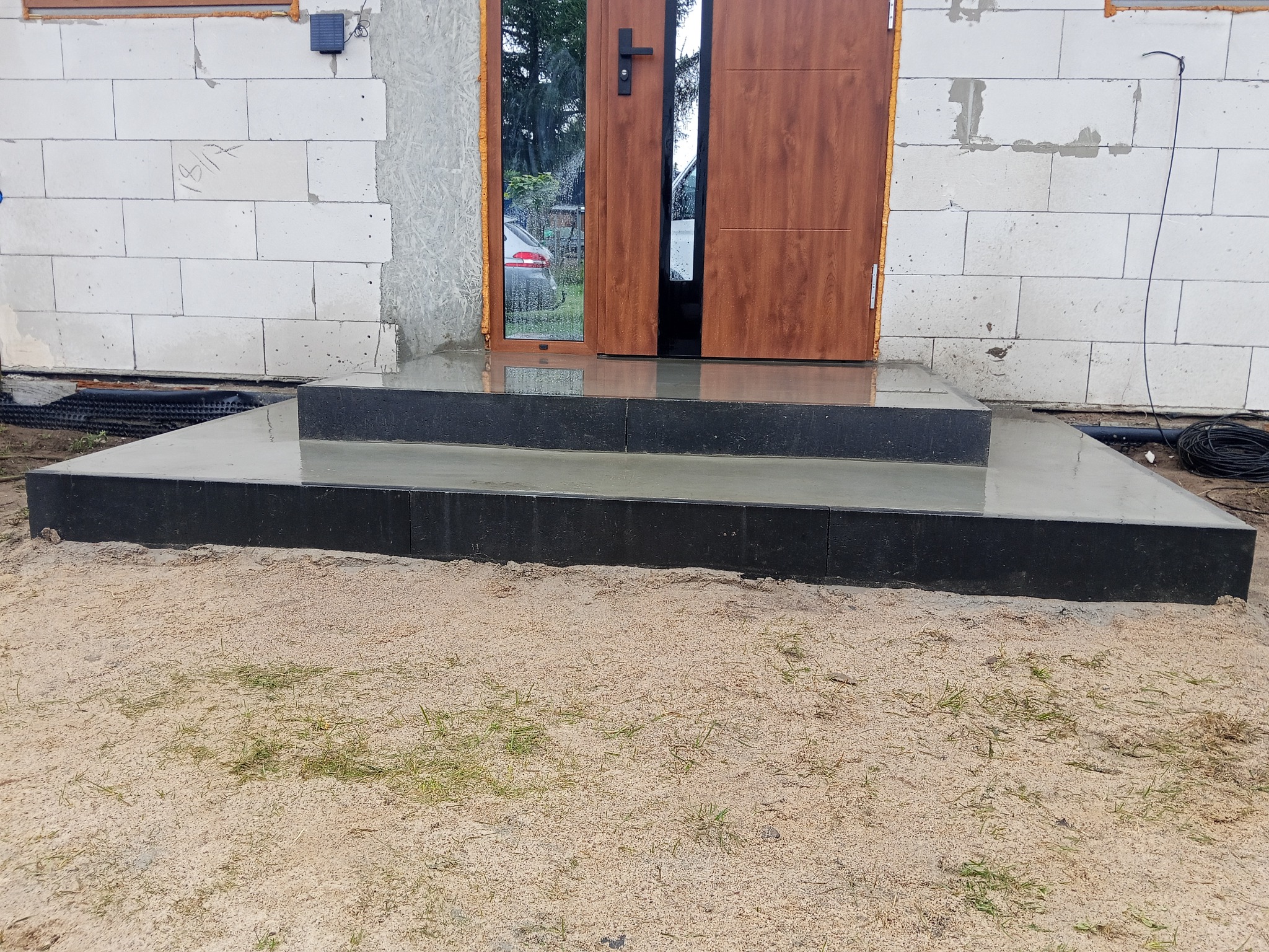 Nowe, dwustopniowe, czarne schody wejściowe z szarego betonu prowadzą do brązowych drzwi w surowym stanie budynku z białego bloczka.