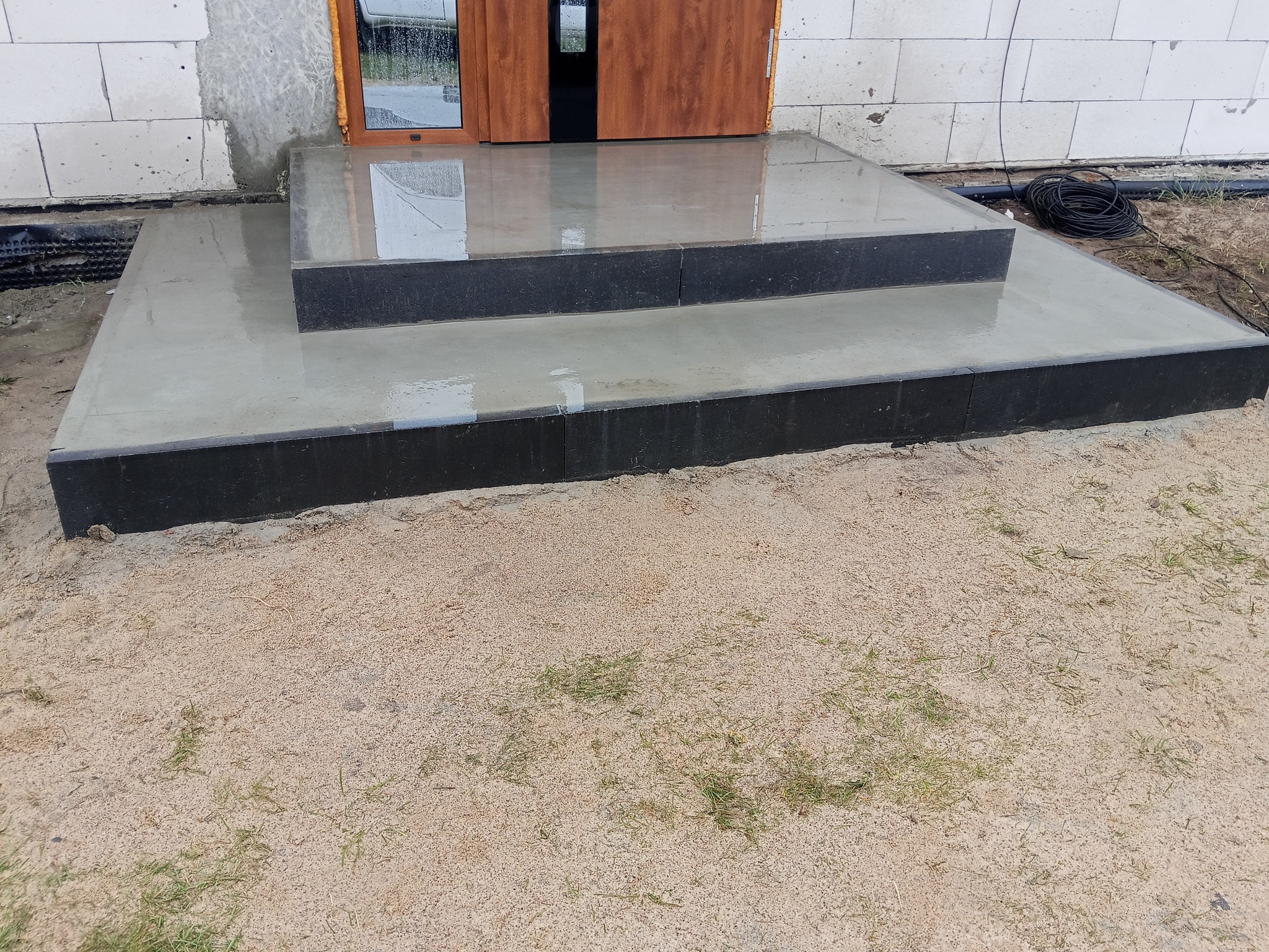 Nowy, dwustopniowy betonowy schodek wejściowy z czarnym obramowaniem, prowadzący do brązowych drzwi w surowym stanie budynku z białego bloczka.