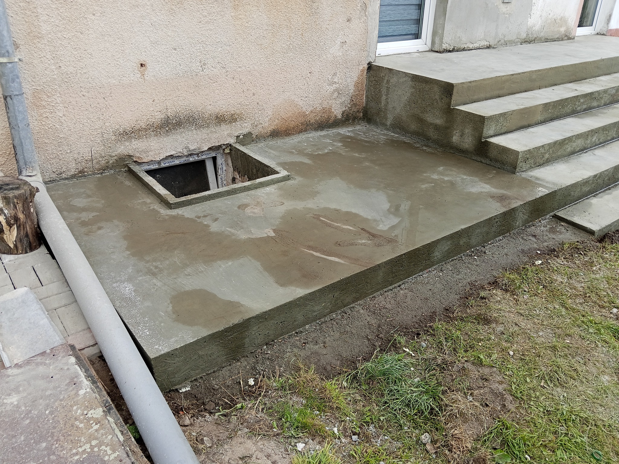 Świeżo wylana, betonowa podstawa z otworem rewizyjnym i schodami wejściowymi do budynku. Widoczne ślady wilgoci na powierzchni betonu.