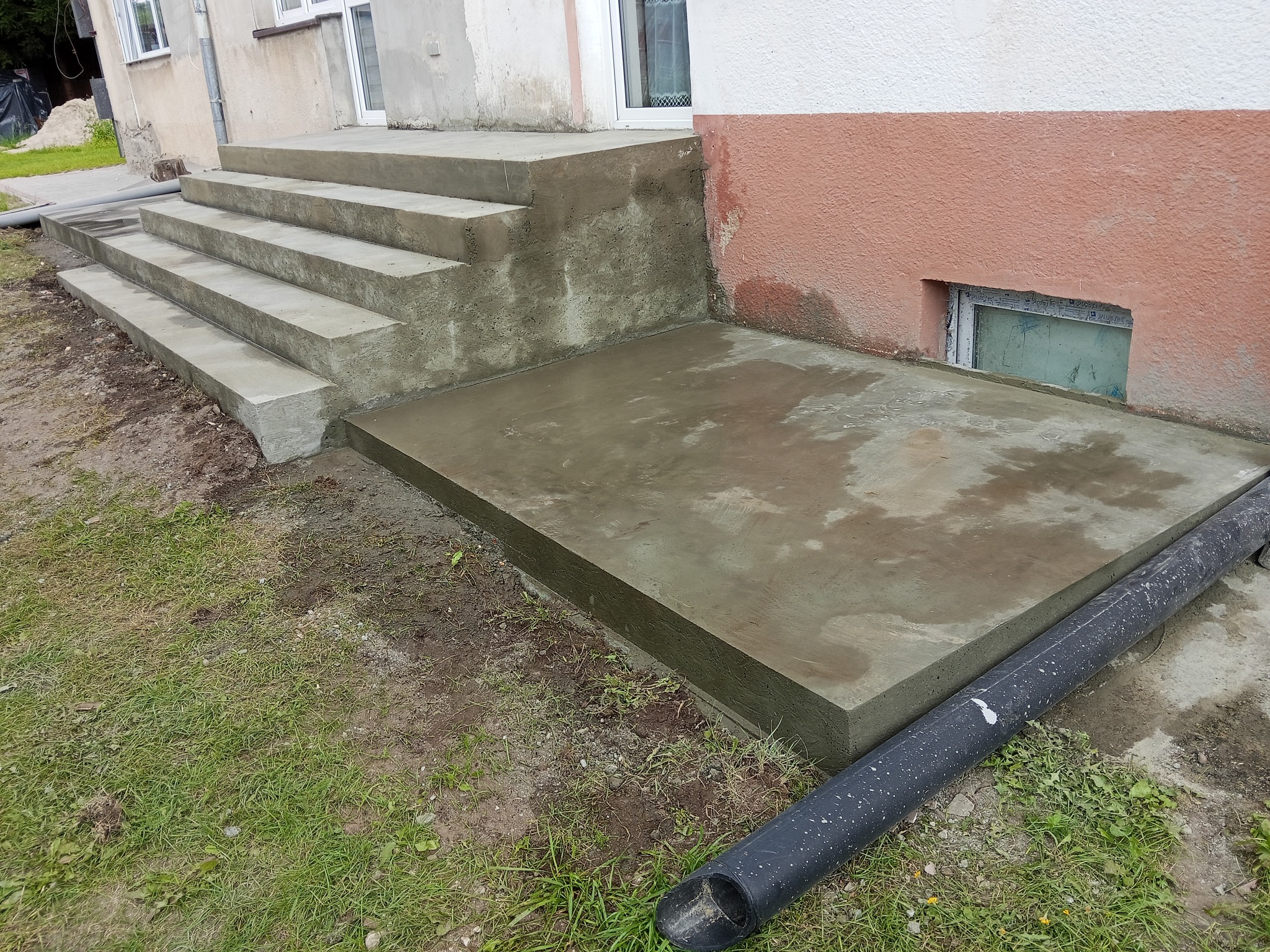 Świeżo wylane, betonowe schody wejściowe do domu z podestem, widoczne ślady wilgoci po procesie wiązania betonu. Rura spustowa wzdłuż podestu.