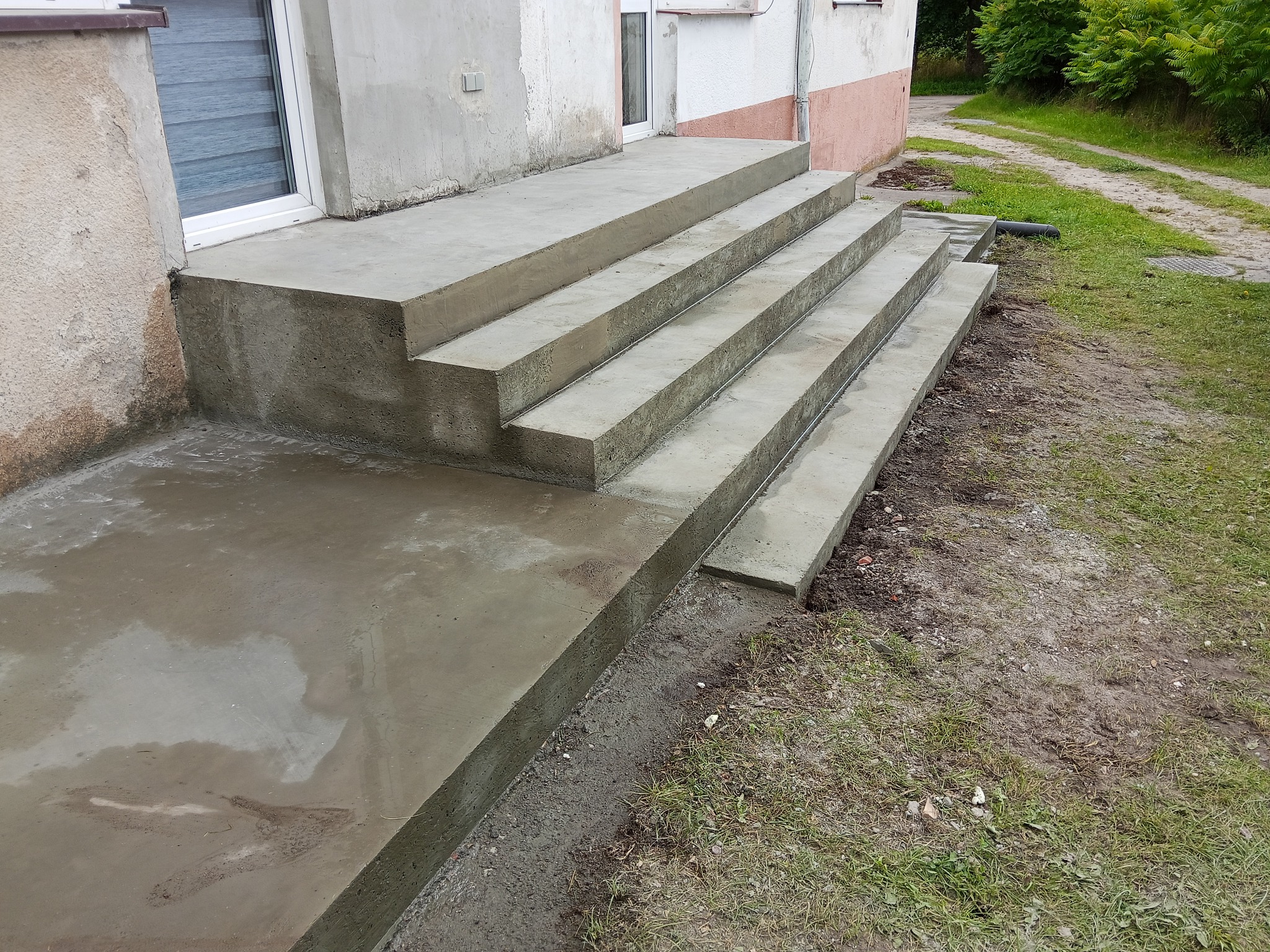 Świeżo wylane, betonowe schody wejściowe do domu jednorodzinnego, z widocznym podestem i czterema stopniami. Tło stanowi trawnik i fragment elewacji budynku.
