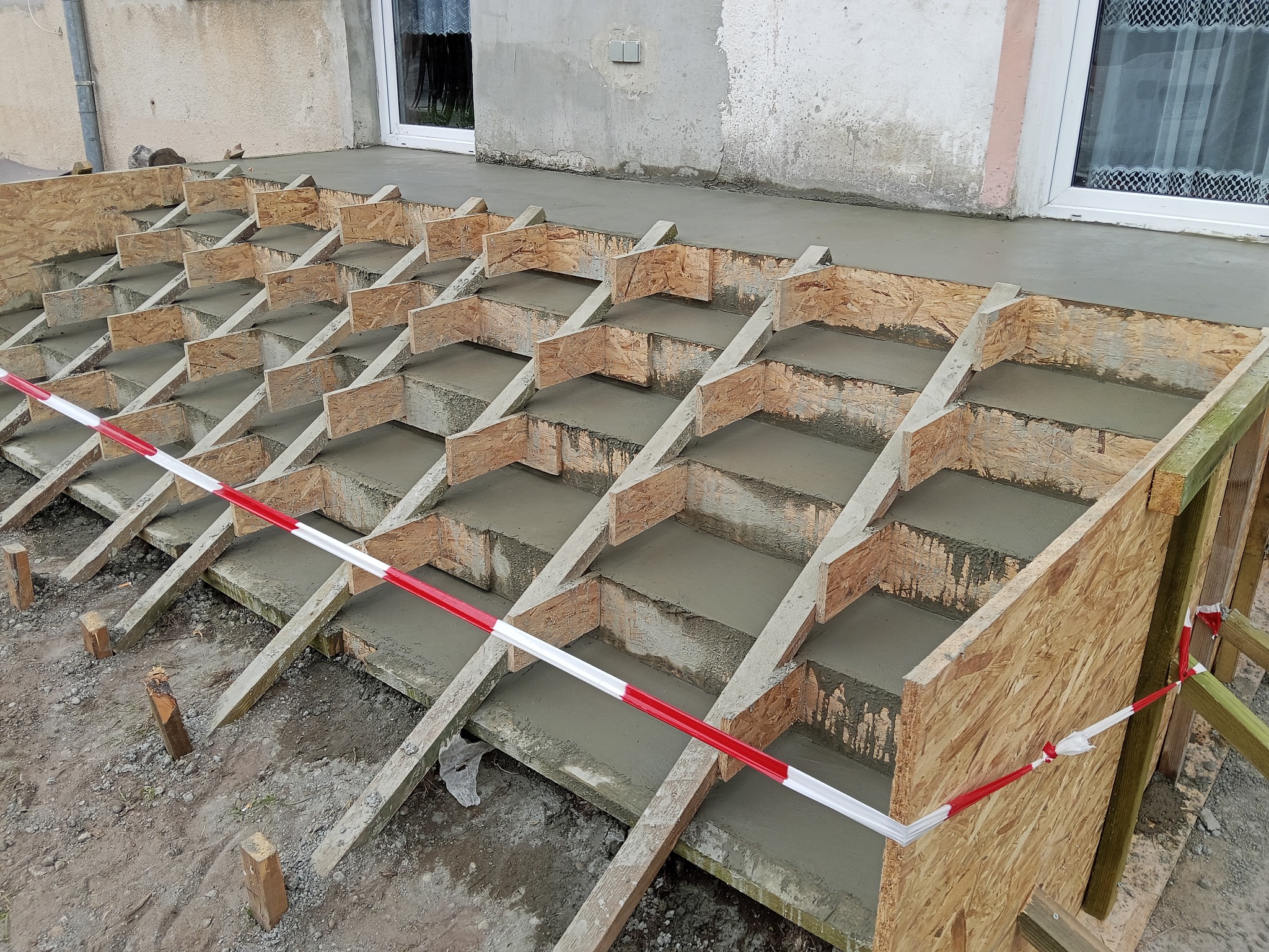 Zalewanie betonem schodów zewnętrznych w drewnianym szalunku, zabezpieczone taśmą ostrzegawczą. Widoczne okna budynku w tle. Etap prac budowlanych.