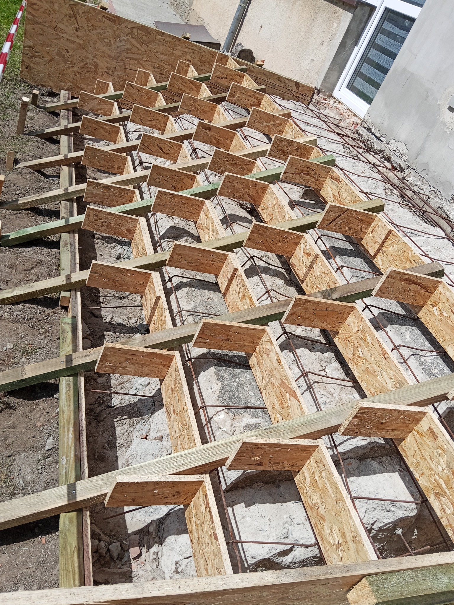 Szkielet drewniany z płyt OSB, przygotowany pod wylewkę fundamentu. Widoczne zbrojenie i fragment ściany budynku. Ujęcie z góry, z bliska.