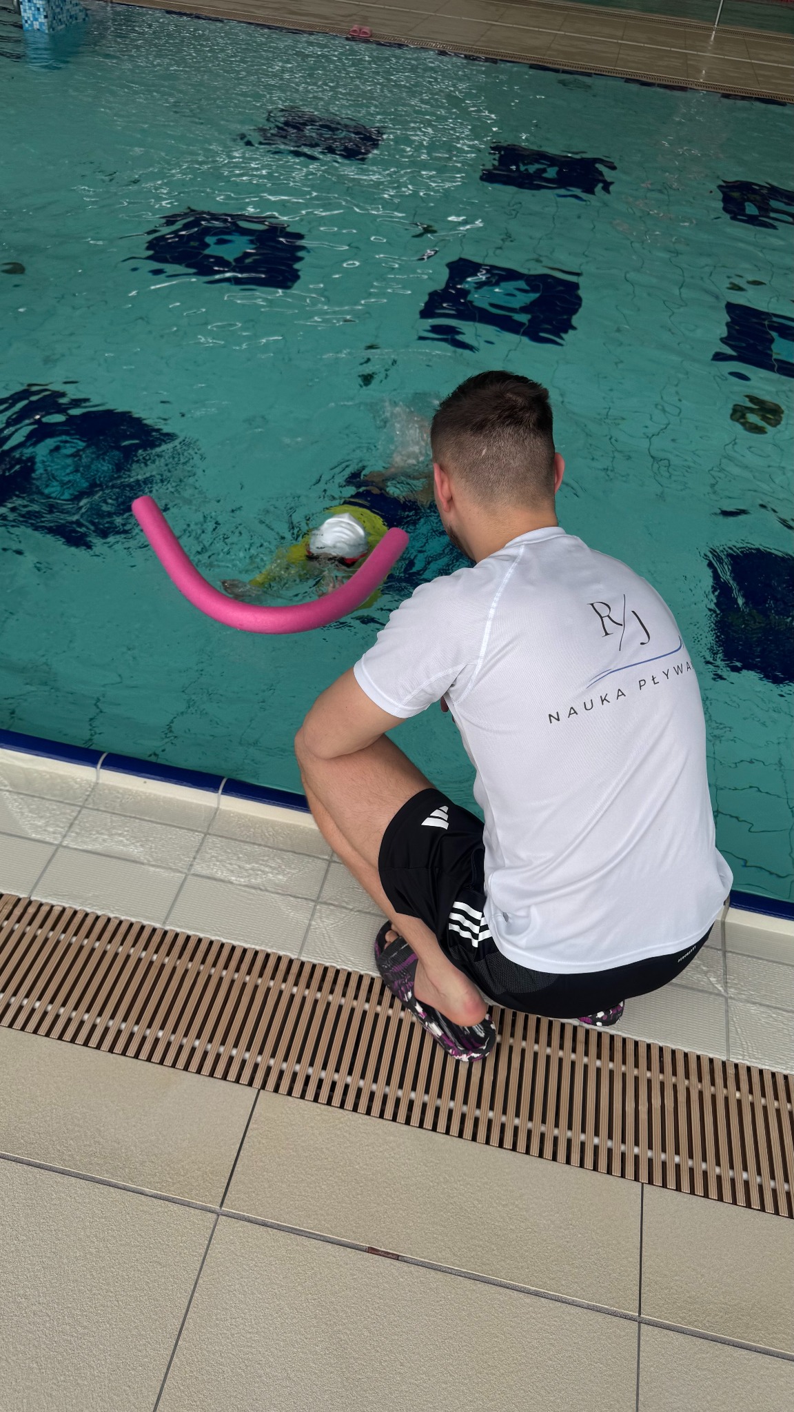 Instruktor w białej koszulce z logo, kuca przy basenie, obserwując dziecko z makaronem do pływania. Widok zza pleców.