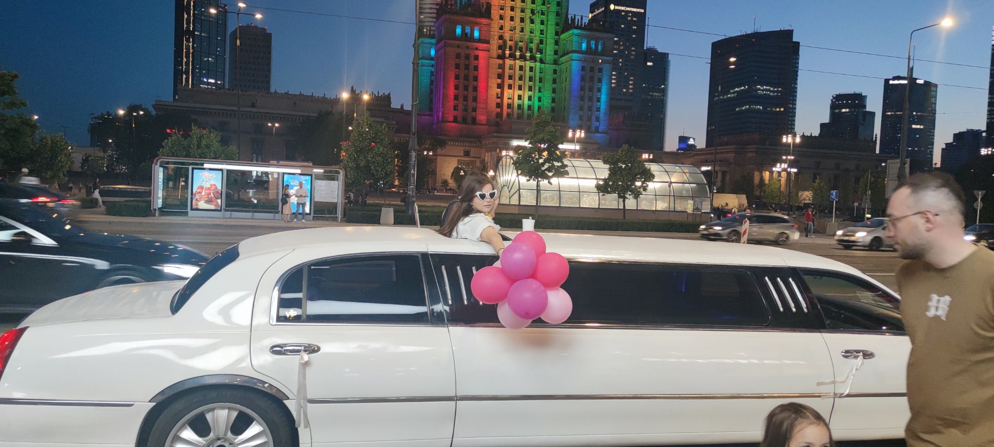 Uśmiechnięta dziewczyna w białej limuzynie z różowymi balonami na tle Pałacu Kultury w Warszawie, wieczorową porą. Okazjonalny wynajem.