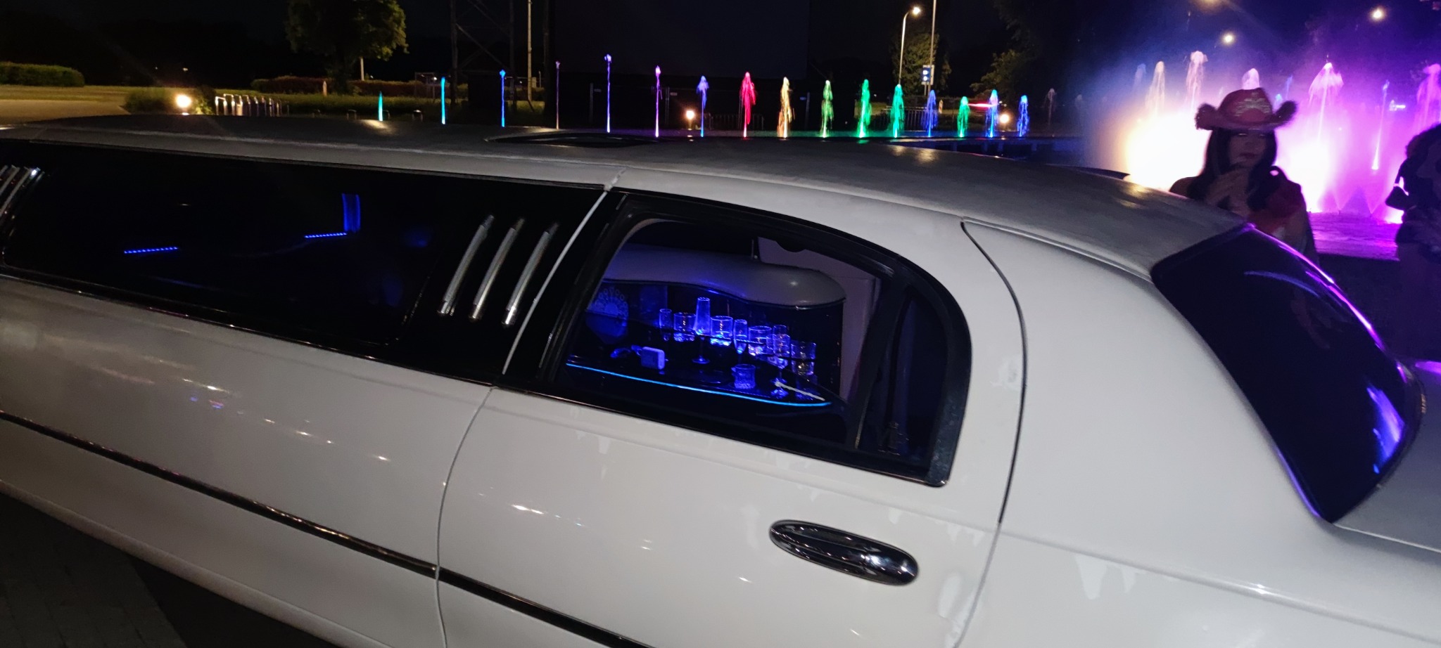 Biała limuzyna na tle fontanny z kolorowym oświetleniem nocą. Wnętrze z barkiem i niebieskim podświetleniem. Częściowo widoczna osoba w kapeluszu kowbojskim.
