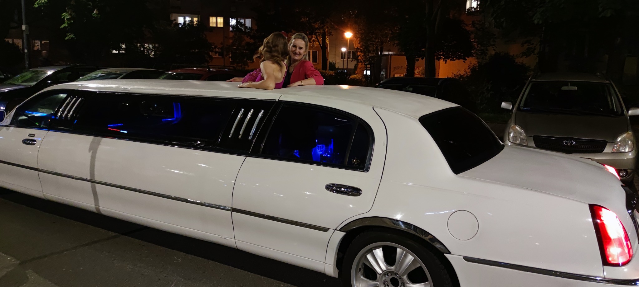 Dwie kobiety w wieczorowych strojach na dachu białej limuzyny z podświetlanym wnętrzem, stojącej na tle nocnego krajobrazu miejskiego.