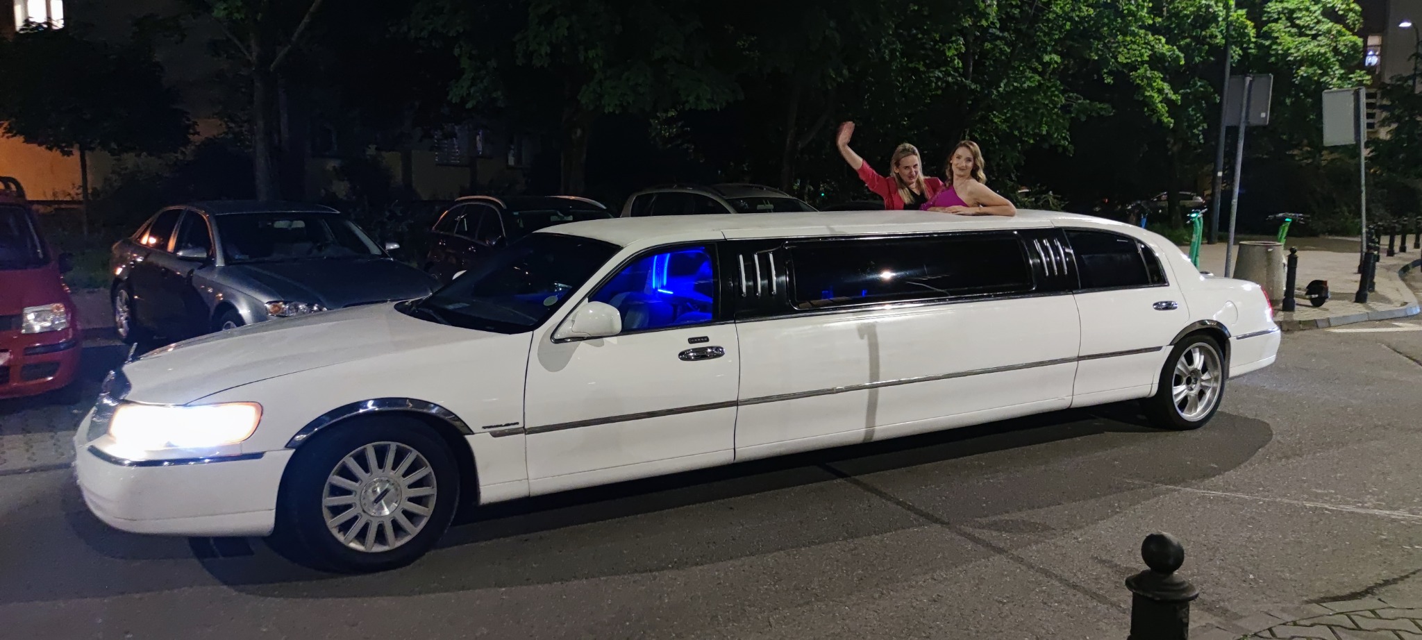 Dwie kobiety w limuzynie Lincoln Town Car, wychylają się przez otwarty dach. Wnętrze oświetlone na niebiesko, nocne otoczenie miejskie.