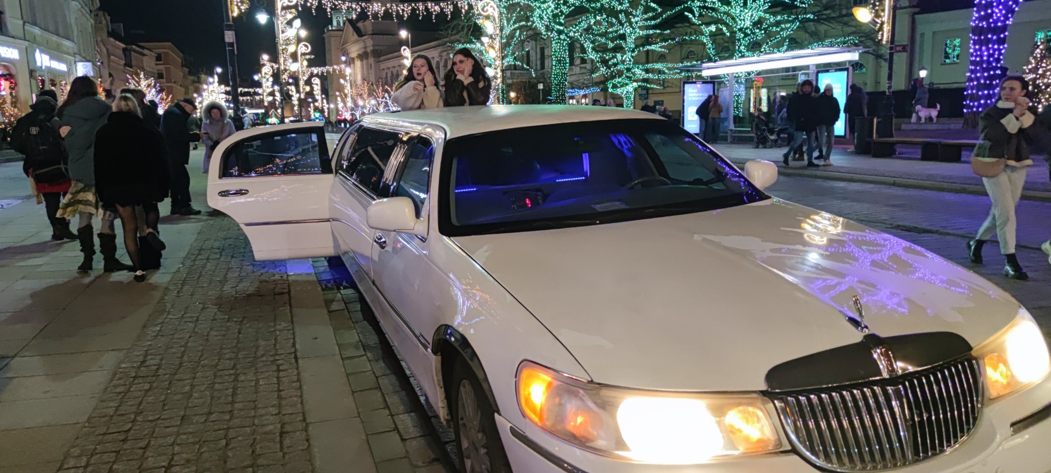 Elegancka biała limuzyna z otwartymi drzwiami na tle świątecznie oświetlonej ulicy nocą. Dwie kobiety wystają z dachu pojazdu, a obok stoją ludzie.