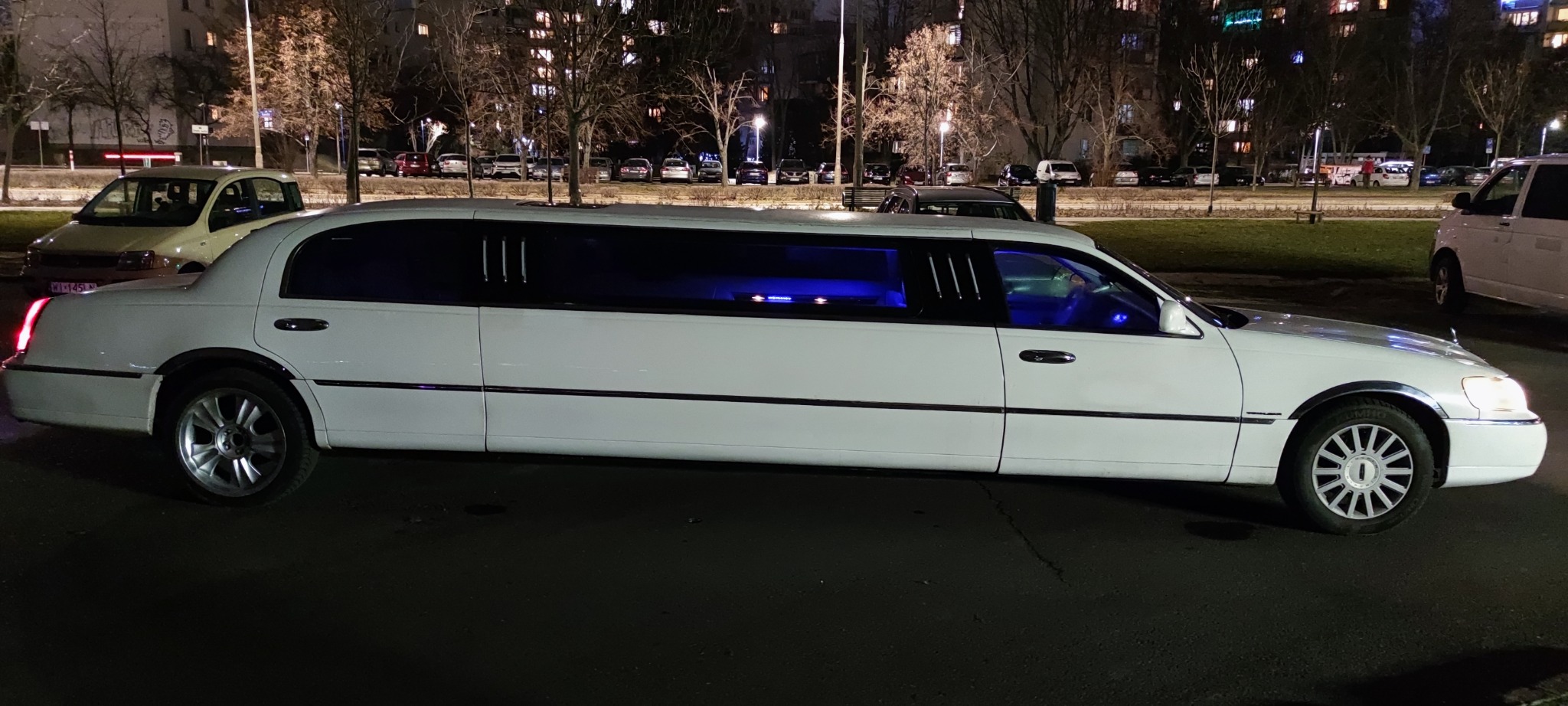 Biała limuzyna Lincoln Town Car z niebieskim oświetleniem wnętrza, zaparkowana nocą na tle bloków mieszkalnych i innych pojazdów.