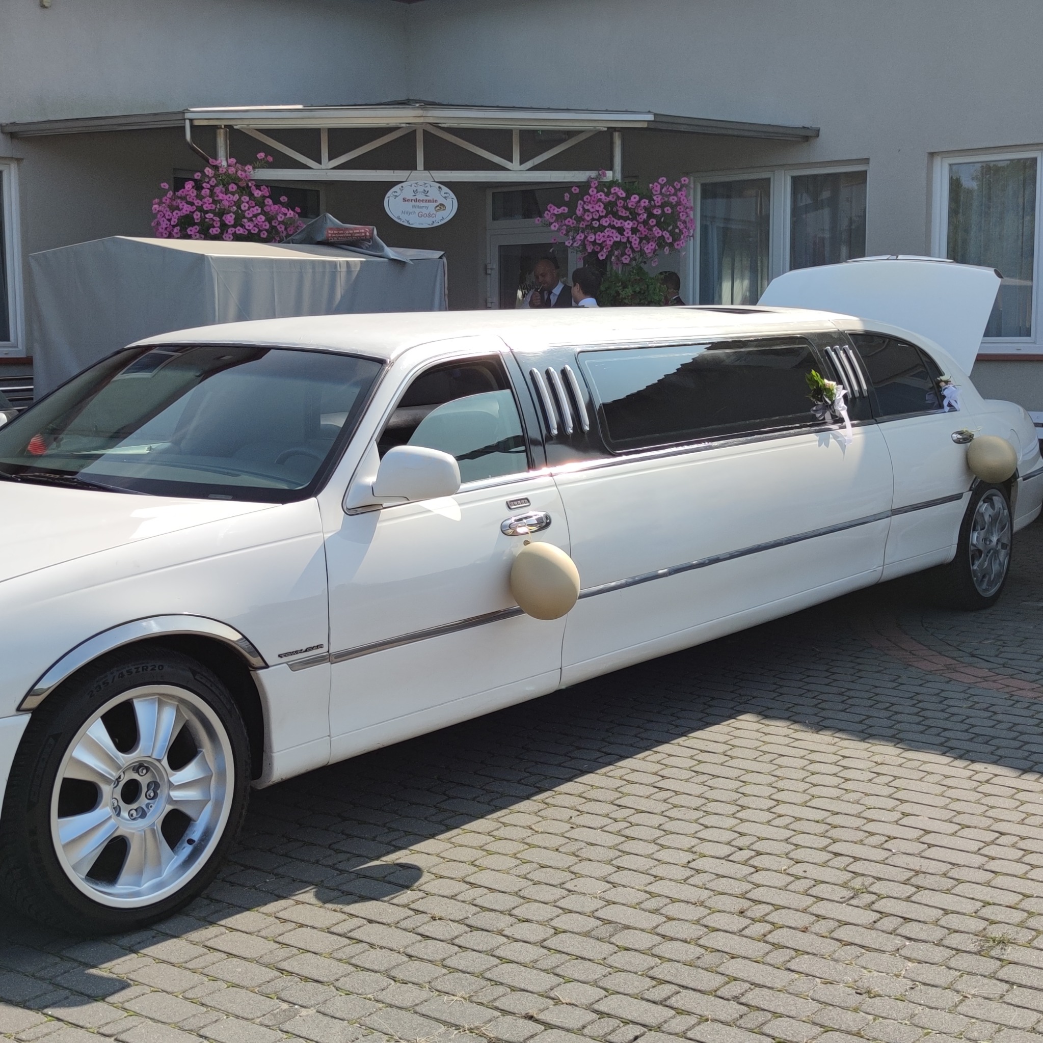 Elegancka, biała limuzyna z otwartym bagażnikiem, udekorowana kremowymi balonami i kwiatami, zaparkowana na brukowanym podjeździe przed budynkiem.