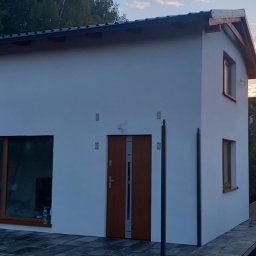 Max Fach Marcin Różek - Biały, dwukondygnacyjny dom z brązowymi oknami i drzwiami, świeżo po elewacji, z widoczną kostką brukową i przyczepką w tle, sfotografowany o zmierzchu.