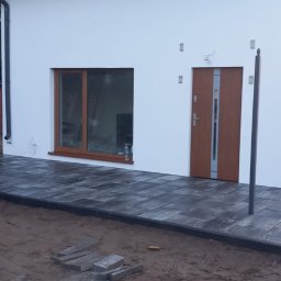 Max Fach Marcin Różek - Nowoczesny dom z jasną elewacją i wybrukowanym tarasem z szarej kostki. Widoczny fragment przyczepy i drewniany budynek w tle.