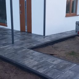 Max Fach Marcin Różek - Nowoczesny dom z białymi ścianami i brukowanym podejściem z szarych płyt. Widoczne okna z drewnianą ramą i brązowe drzwi wejściowe.