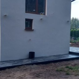 Max Fach Marcin Różek - Wykończona elewacja domu z dwoma oknami i czarną rurą spustową przy tarasie z kostki. Widoczne przewody elektryczne przy górnym oknie.