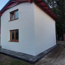 Max Fach Marcin Różek - Biała elewacja domu z brązowymi oknami i czerwonym dachem, widok z boku na tle zieleni, przyczepa i kostka brukowa w otoczeniu.