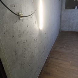 Max Fach Marcin Różek - Surowa ściana z widoczną fakturą po szpachlowaniu, oświetlona pionową lampą, z fragmentem brązowej podłogi z płytek i gniazdkiem elektrycznym.