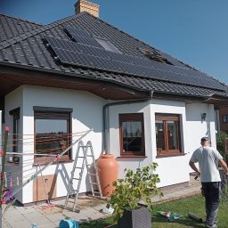 Max Fach Marcin Różek - Biały dom z ciemną dachówką i panelami słonecznymi. Widoczna drabina, zbiornik na deszczówkę i osoba z przewodami, co sugeruje instalację paneli.
