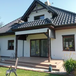 Biała elewacja domu z ciemną dachówką i drewnianymi elementami. Widoczny taras, okna z brązowymi ramami i zieleń w ogrodzie.