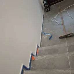 Świeżo pomalowana ściana na schodach zabezpieczona taśmą malarską, widoczny fragment podłogi z narzędziami malarskimi i radio.