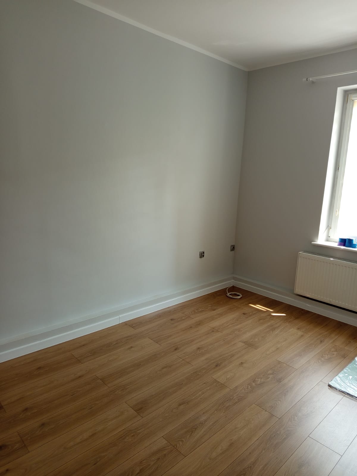 Pokój po remoncie: szare ściany, drewniana podłoga, białe listwy przypodłogowe i okno z grzejnikiem. Widoczne gniazdka elektryczne i kabel.  Jasne, dzienne światło.