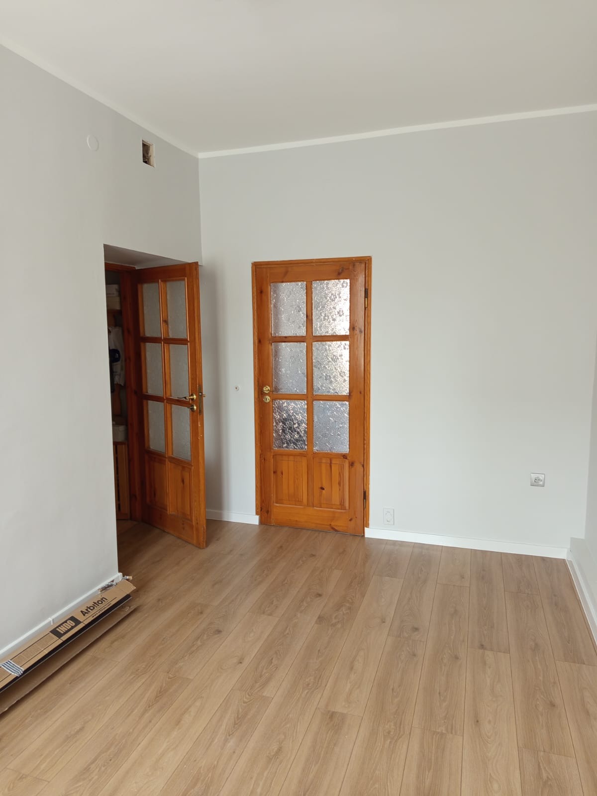 Jasne wnętrze po remoncie: drewniana podłoga, dwoje przeszklonych, drewnianych drzwi, ściany pomalowane na szaro. Widoczne listwy przypodłogowe i karton z panelami.