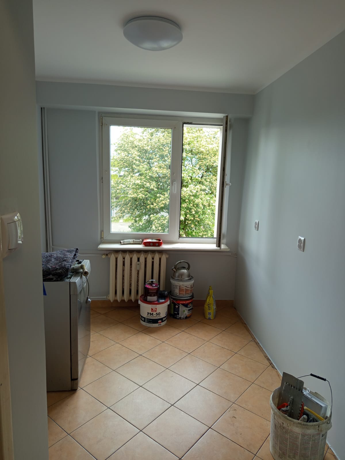 Wnętrze pokoju w trakcie odświeżania: widoczne puszki z farbą PM-50, narzędzia malarskie w wiadrze, grzejnik i okno z widokiem na zieleń. Ściany w kolorze jasnego błękitu.