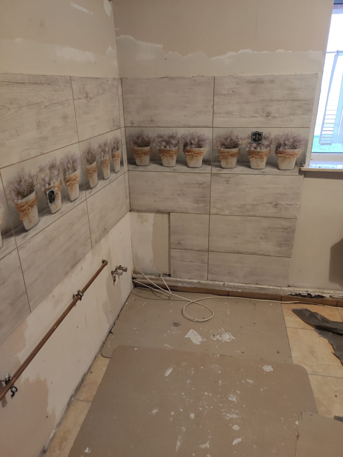Wnętrze w trakcie remontu: częściowo wyłożona płytkami ściana z motywem kwiatów w doniczkach, widoczne rury i przewody, podłoga z resztkami zaprawy.