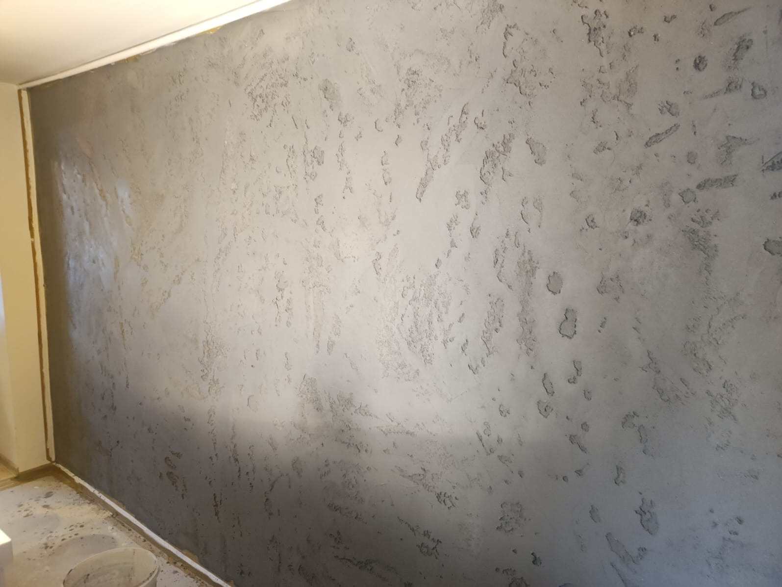 Szara ściana z wyraźną, nieregularną fakturą, lekko połyskująca w górnej części. Widoczny fragment podłogi i narożnik pokoju. Dekoracyjne wykończenie ściany.