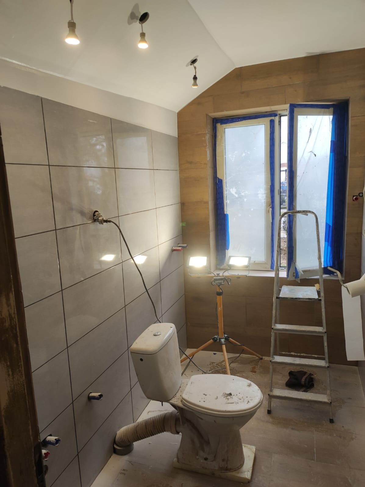 Łazienka w trakcie remontu: płytki na ścianach, zabezpieczone okno, drabina, przenośne oświetlenie. Widoczne ślady prac wykończeniowych.
