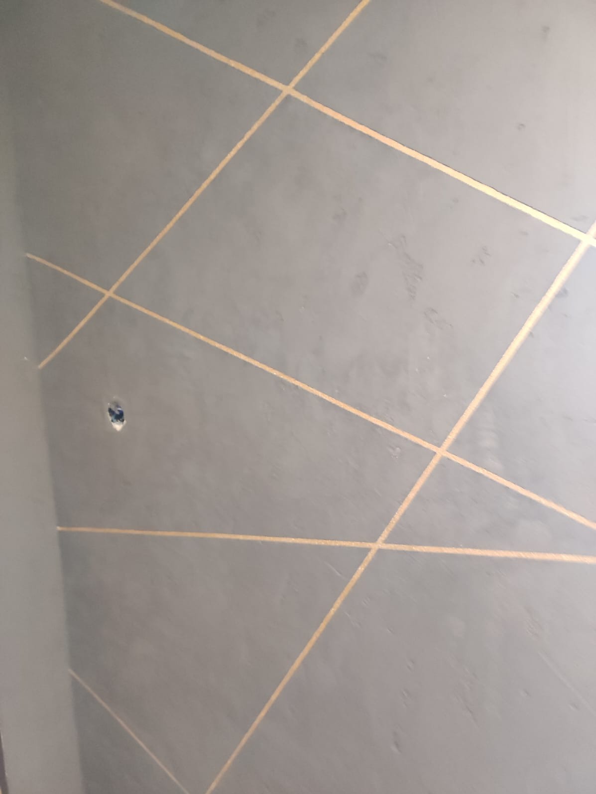 Szara ściana z dekoracyjnymi, złotymi liniami tworzącymi geometryczny wzór. Widoczne ślady szpachlowania i otwór po gniazdku.