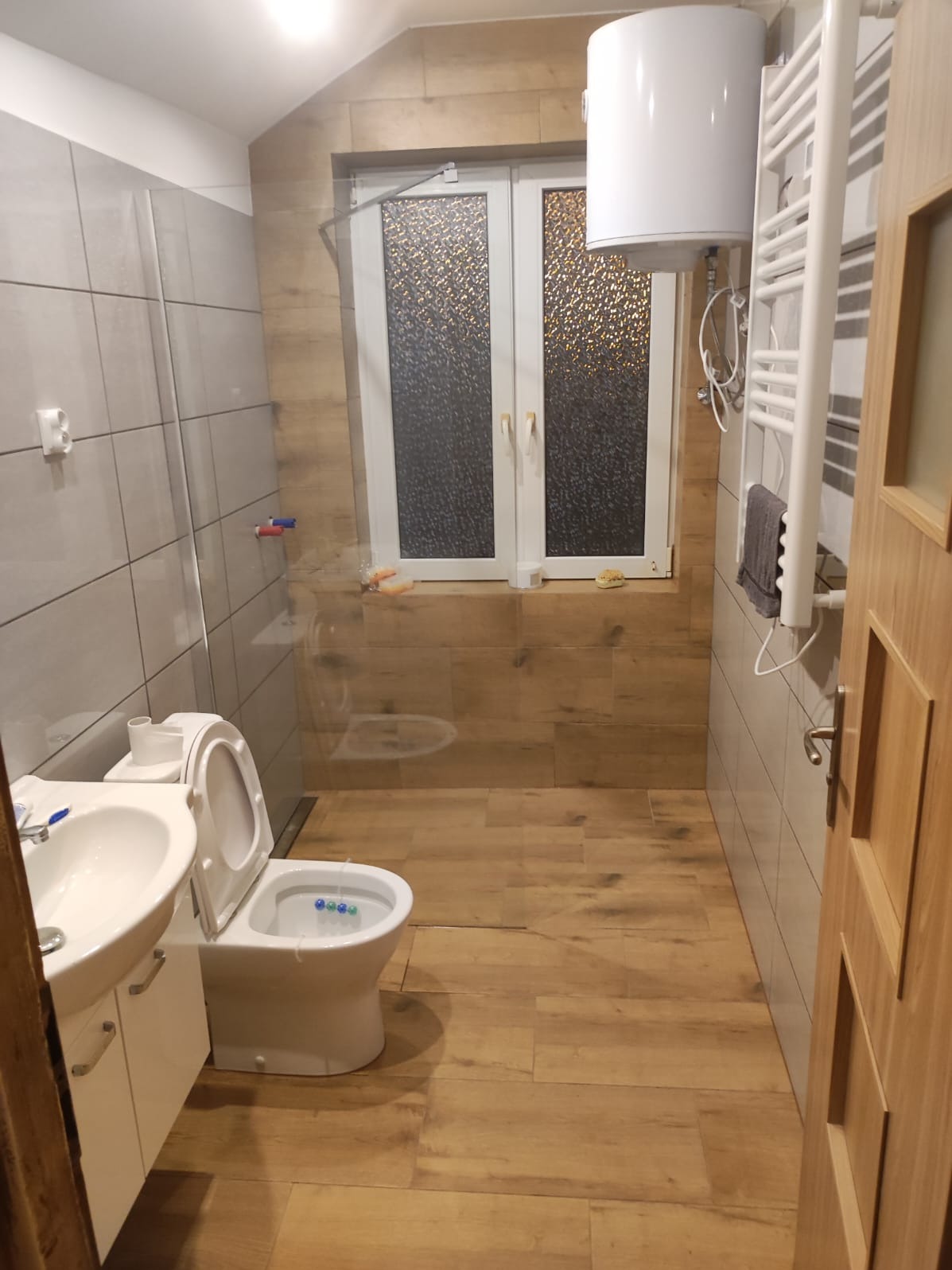 Jasna łazienka po remoncie z drewnopodobnymi płytkami na podłodze i ścianach, białą armaturą i grzejnikiem drabinkowym. Widoczne okno z ozdobną szybą.